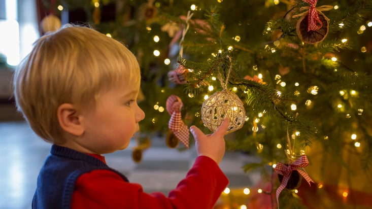 A young boy wearing a blue knitted tank top points at a natural woven bauble on a Christmas tree glowing with fairy lights